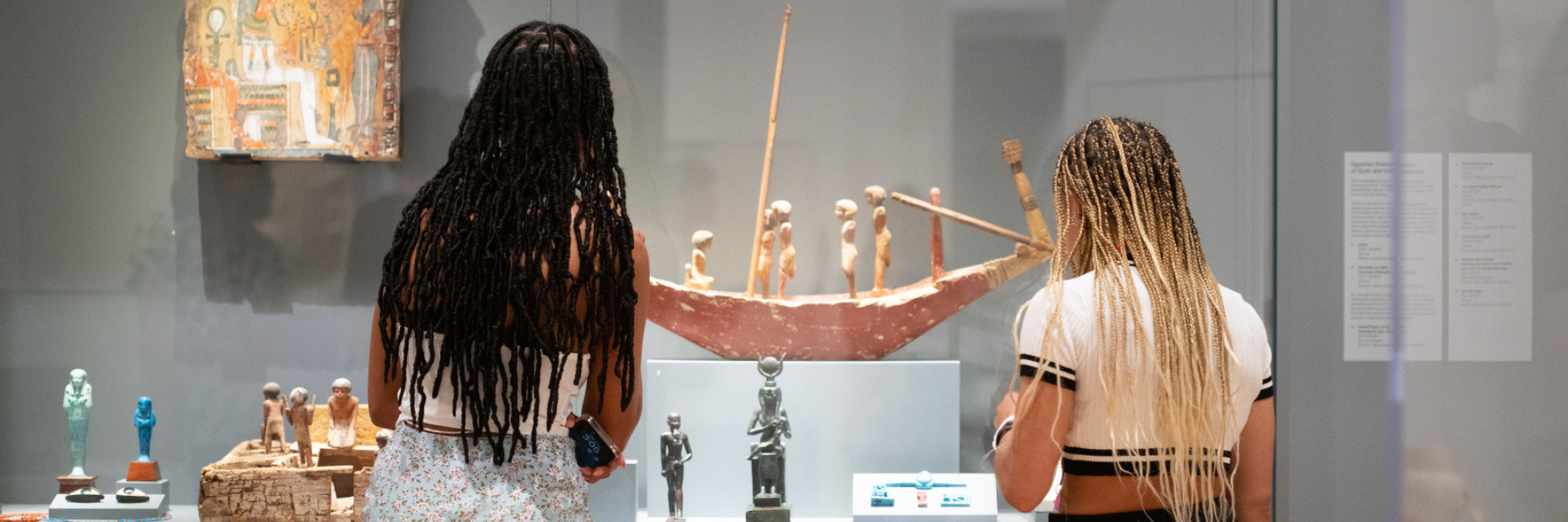 Two people stand peering into a museum display case facing away from the viewer. They both have medium complexion skin, and waist length braids. The person to the left has dark hair, and is wearing a white shirt with a light blue and white skirt. The person to the right has blond and dark hair, and is wearing a white shirt with a black border and black pants. 